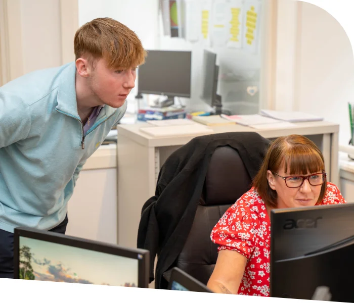 A TMQ employee in a light blue top leans over to look at a computer screen being used by another TMQ accountant in a red floral top sitting at a desk, discussing accountancy services in an office environment.