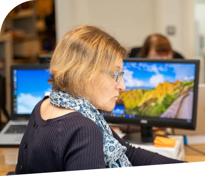 Susanne Robertson with short hair, glasses, and a scarf works at a desk with two computer monitors displaying different background images, likely providing accountancy support services. Another person is visible in the background blurred.