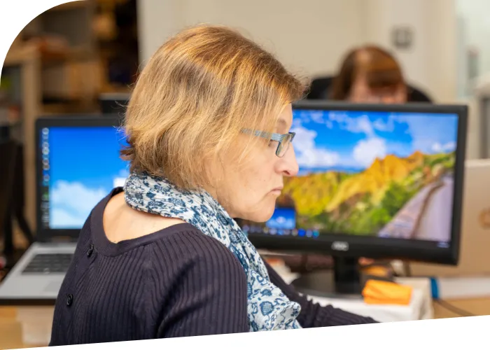 Susanne Robertson with short hair, glasses, and a scarf works at a desk with two computer monitors displaying different background images, likely providing accountancy support services. Another person is visible in the background blurred.