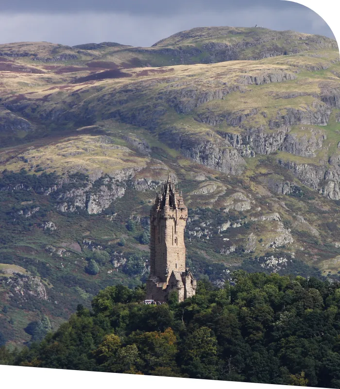 A tall, stone tower stands among lush greenery on a hill with a backdrop of rolling, rocky mountains under an overcast sky, guiding the diligent accountants of Stirling through their scenic landscape.