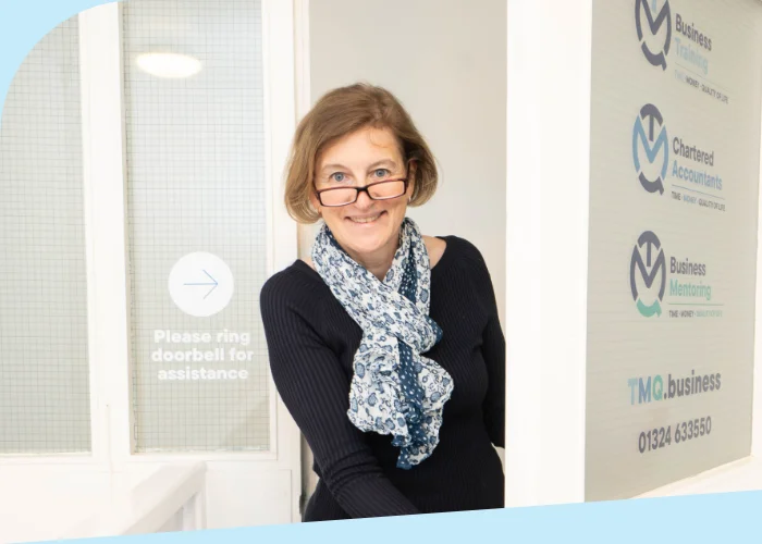 Susanne Robertson opens a door in an office hallway at Falkirk Accountants. She is wearing glasses and a patterned scarf, smiling at the camera. The door has company logos and text on it, adding to the welcoming atmosphere.