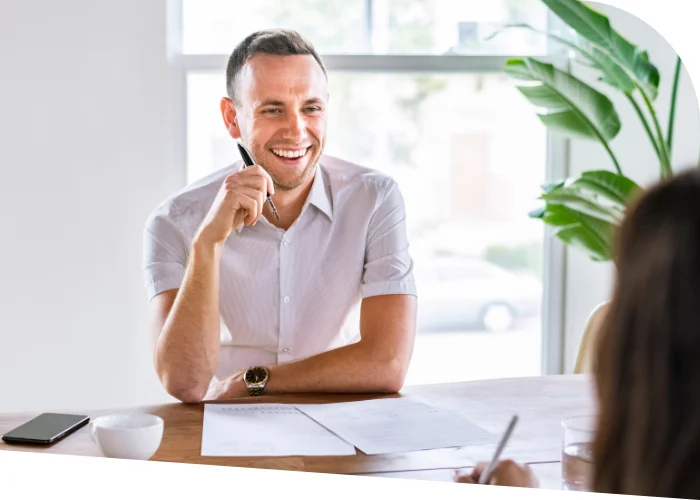 A man in a white shirt is smiling while holding a pen and sitting at a desk with papers. A smartphone, a bowl, and a plant are on the table. A person with long hair is seated across from him, likely discussing their transformative pricing strategy.